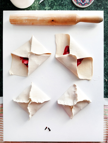Photo recipe and sequence for preparing square baked pies envelopes from flaky puff pastry stuffed with apples, cherries, orange and liquor fillings on a white board and kitchen table.