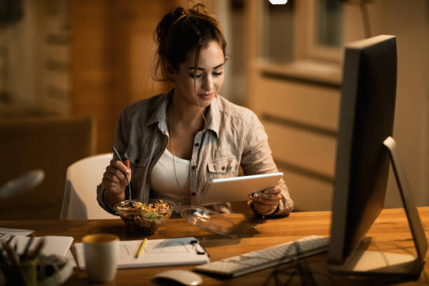 istock Young woman using touchpad while eating salad at night at home. 1192151711