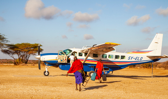 Masai guides carrying luggage to safari plane on the dry savannah close to amboseli Kenya. Clear sky with a few fluffy white clouds