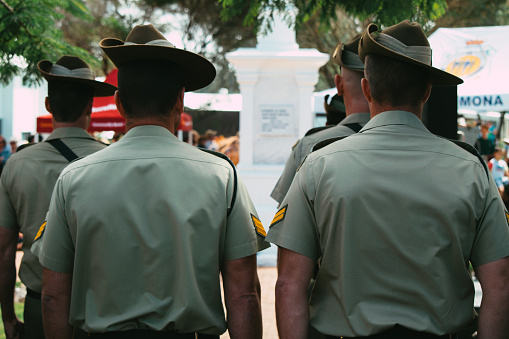 A rear-facing shot of a section of Australian Army soldiers, a sergeant and corporal, wearing traditional ceremonial uniforms, with Slouch Hat and slung F88 Austeyr Rifles (black weapon slings are visible) standing in the shade whilst waiting to take their part in the ANZAC Day remembrance ceremony at the Cooroy cenotaph, as the official Honour Guard.