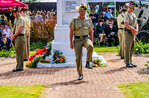 A section of Australian Army soldiers wearing ceremonial uniforms, with Slouch Hat and slung F88 Austeyr Rifles, move into position with traditional drill movements during the ANZAC Day remembrance ceremony at the Cooroy cenotaph. The soldiers with heads bowed are the official Honour Guard for the service. Members of the crowd are visible in the background. This image could be used to serve as a metaphor for reflection, military service, teamwork, memory or national pride.