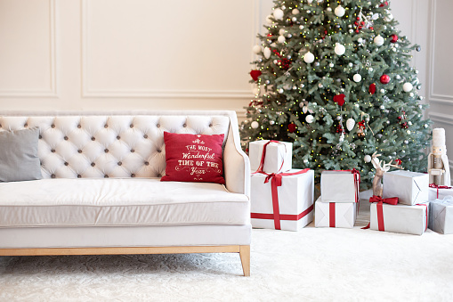 Luxury living room interior with sofa decorated chic Christmas tree, gifts and pillows. Classic interior in red shades. Christmas at home. beige sofa on background of Christmas tree. \nNew Year decor