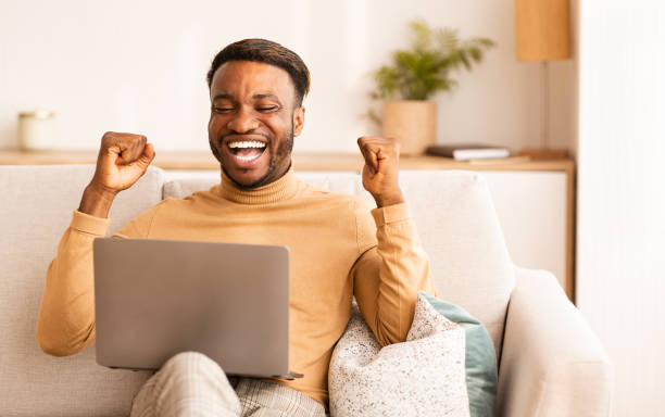 istock Black Man Using Laptop Gesturing Yes Sitting On Couch Indoor 1192256070