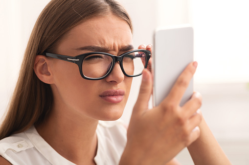 Poor Eyesight. Girl Squinting Eyes Looking At Smartphone Screen Through Eyeglasses Sitting Indoor. Selective Focus