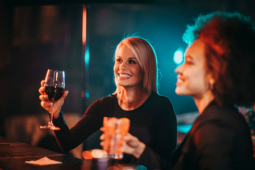Two women enjoying their drinks while sitting at a bar table in a night club. One of them is having a glass of wine and the other a cocktail.