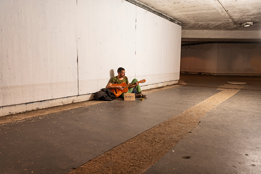 Photo of Homeless Man who is Sitting in the Train Station, Subway Station, Tunnel or Underground and Playing Guitar with Hungry Sign and Donation Box. Desperate Mature Man is Feeling Abandoned and Lost in Depression and Sitting on Ground Street While Suffering Emotional Pain.