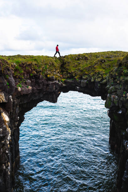 Tourist walks over a natural rock bridge in Arnarstapi, Iceland Tourist walks over a natural rock bridge connecting basalt cliffs in Arnarstapi, Iceland, with Atlantic Ocean in the background. spectacular view stock pictures, royalty-free photos & images