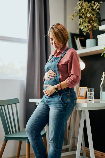 Attractive proud caucasian future pregnant mother leaning on desk at home office and touching belly.