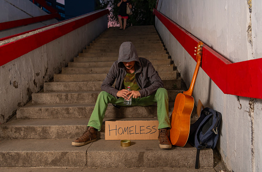 Photo from Above of Homeless Person who is Sitting on the Stairways While Holding his Acoustic Guitar and Drinking an Alcohol. Sad and Desperate Young Man or Beggar is Drinking Vodka While Asking for Money Donation. Poverty and Social Issue Concept.