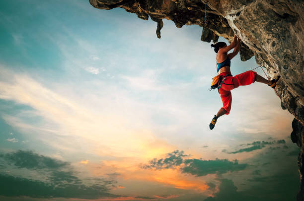 日没の空の背景を持つ張り出した崖の岩に登るアスレチックな女性 - overhanging ストックフォトと画像
