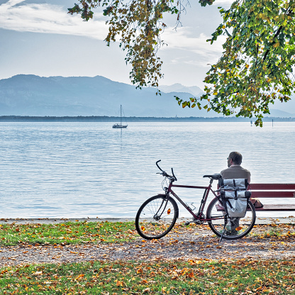 Lindau, Germany - October 27, 2019: A man sitting on a bench next to a bicycle lookig at the Bodensee and Alps in the Toscana Park, Lindau, Germany.