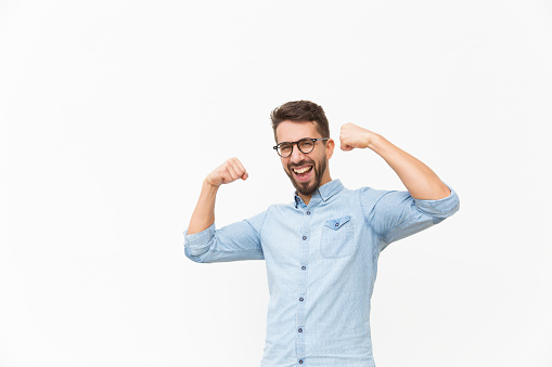 Happy joyful guy making hand winner gesture. Handsome young man in casual shirt and glasses standing isolated over white background. Success concept