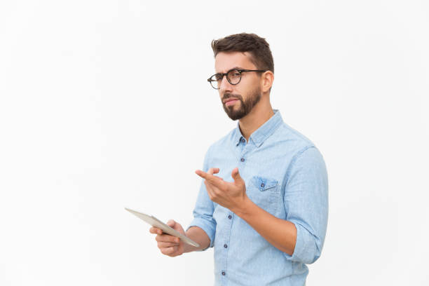 Pensive tablet user looking at camera and pointing finger away Pensive tablet user looking at camera and pointing finger away. Handsome young man in casual shirt and glasses standing isolated over white background. Advertising concept suspicion stock pictures, royalty-free photos & images
