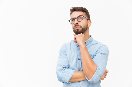 Pensive customer thinking over special offer, touching chin, looking up. Handsome young man in casual shirt and glasses standing isolated over white background. Advertising concept