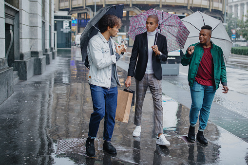 Three young men with shopping bags walking in the rain holding umbrellas