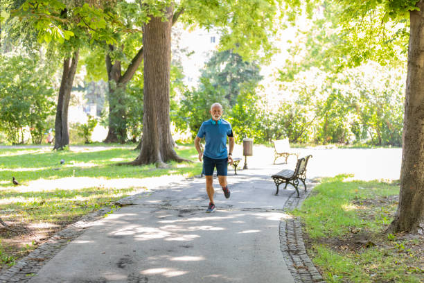 Mature Man with Grey Hair and Beard is Jogging in the City Park and Listening to Music. Handsome Older Man with Wearable Computer and Headphones is Taking Morning Run in the Sports Track in Public Park.