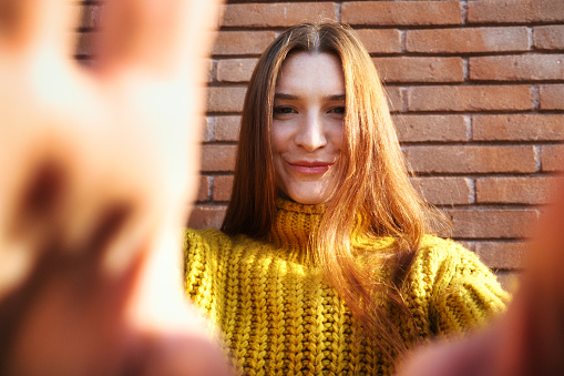 Shy Young Redhead Woman Covering Camera Lens With Hands And Smiling Candid Against A Brick Red Wall.