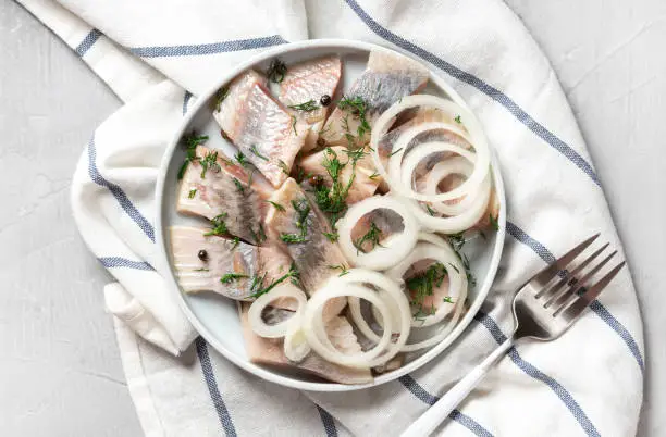 Pieces of pickled herring with onions, dill and pepper in a ceramic plate on a light gray background, top view, copy space.