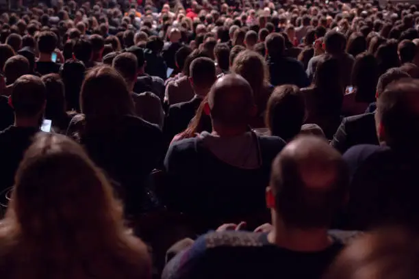 Photo of In the dark hall there is a view from the back of a crowd of hundreds of people sitting and watching the screen in a movie theater or performing on stage.