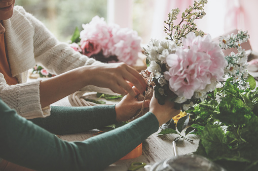 Side view of two unrecognizable young women sitting at the table and tying a piece of twine on a beautiful flower arrangement they are making.