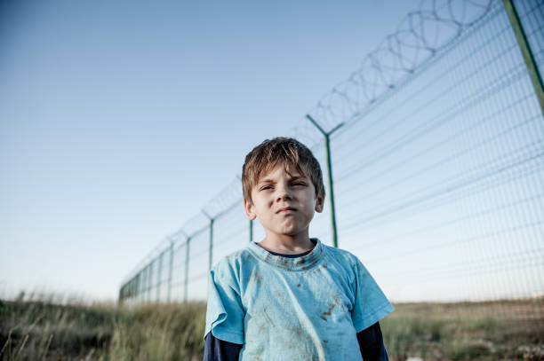 portrait of victim of war conflict poor orphan refugee prisoner migrant child in dirty clothes with sad face near state border with fence with barbed razor wire and blue sky portrait of poor orphan refugee prisoner migrant child in dirty clothes with sad face near state border with high fence with barbed razor wire war zone stock pictures, royalty-free photos & images