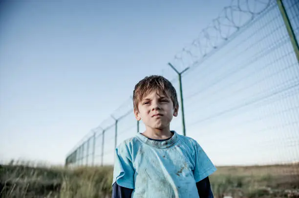 portrait of poor orphan refugee prisoner migrant child in dirty clothes with sad face near state border with high fence with barbed razor wire