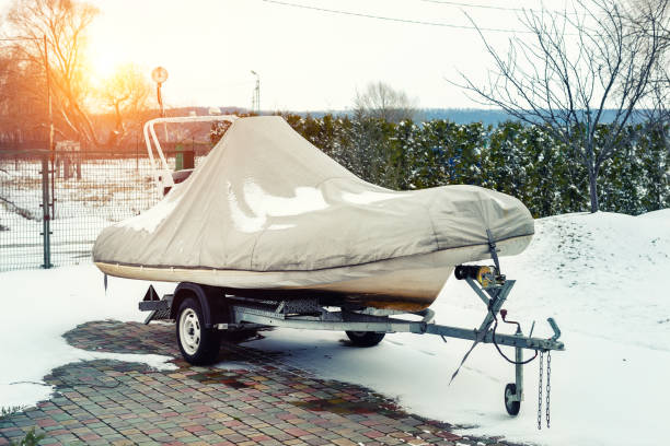 裏庭の冬期の季節の貯蔵のためのトレーラーの上に立ってカバーに包まれた膨脹可能な贅沢な釣りモーターボート。駐車場で冬に収縮した容器 - ラップドレス ストックフォトと画像