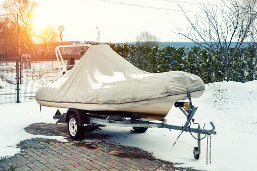 Inflatable luxury fishing motorboat wrapped in cover standing over trailer for winter period seasonal storage at backyard. Shrink-wrapped vessel winterized on parking.