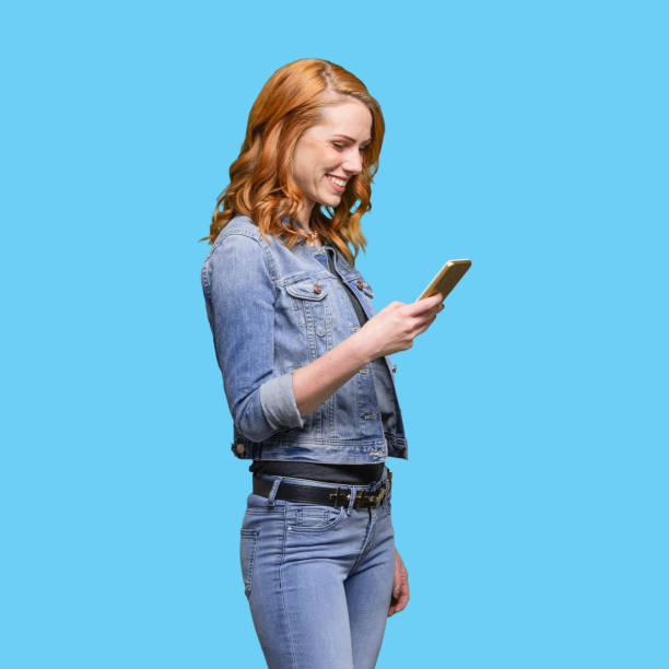 one person / waist up / side view / profile view / looking down of 20-29 years old adult beautiful redhead caucasian female / young women standing wearing jeans / pants / double denim / denim jacket who is smiling / happy / cheerful / laughing - 20 25 years profile female young adult imagens e fotografias de stock