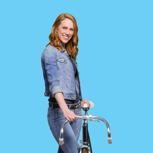 one person / waist up / side view / profile view / looking at camera of 20-29 years old adult beautiful redhead / long hair caucasian female / young women standing wearing jeans / pants / double denim / denim jacket / cool attitude and using bicycle - 20 25 years profile female young adult imagens e fotografias de stock