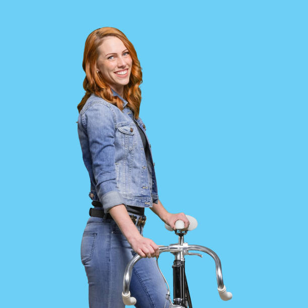 one person / waist up / side view / profile view / looking at camera of 20-29 years old adult beautiful redhead / long hair caucasian female / young women standing wearing jeans / pants / double denim / denim jacket / cool attitude and using bicycle - 20 25 years profile female young adult imagens e fotografias de stock