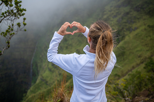 Young woman making heart shape finger frame on waterfall and mountain in Hawaii