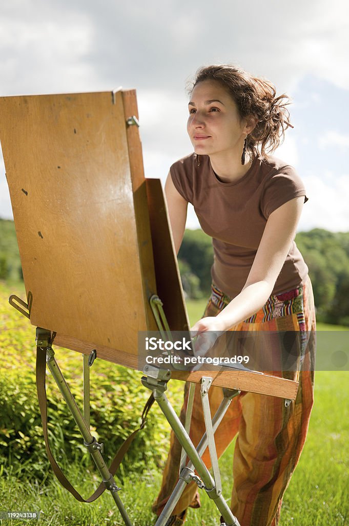 Young woman looking at a painting easel outside in a field Pretty woman is painting. Open air session. Outdoors Stock Photo