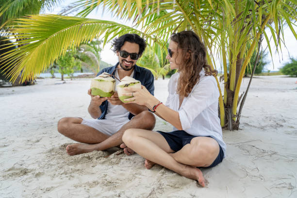 istock A beautiful young couple joyfully sits on white sand under a palm tree with coconuts in their hands on the seashore under a green palm tree. Honeymoon Travel and Vacation on the Tropical Ocean 1192433834
