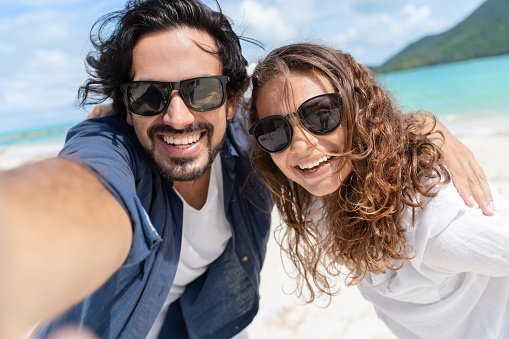 Beach vacation couple taking selfie photograph using smartphone relaxing and having fun holding smart phone camera. Young beautiful multicultural Latin Caucasian couple on summer beach.