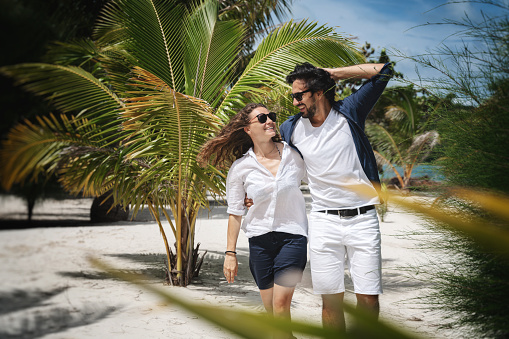 Beautiful young couple joyfully walks on the white sand on the seashore between green palm trees. Honeymoon Travel and Vacation on the Tropical Ocean.