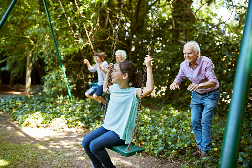 Active senior Argentine grandparents pushing preteen male and female grandchildren on swings in family backyard.