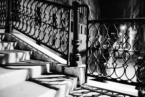 Detail in black and white of the iron banister of a bridge in Venice at night. The shadow of the iron work draws lines on the white stone stairs, the street lights and canal are in the background