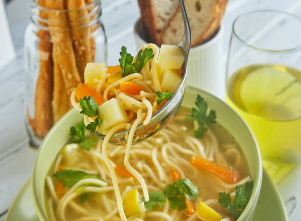 italian soup with spaghetti, carrots, lemon, parsley and pieces of chicken in a green plate, on a table with napkins, spoons of bread and a drink in a glass - basil tomato soup food and drink imagens e fotografias de stock