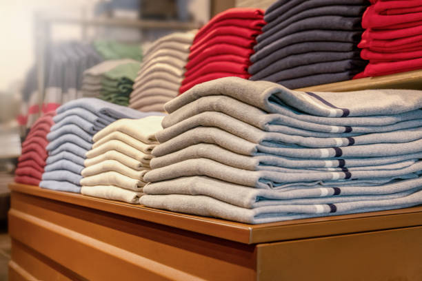 Wide angle color image depicting piles of hundreds of men's winter sweaters stacked on wooden shelves inside a store. The jumpers are in different colors including green, blue and grey. Room for copy space.