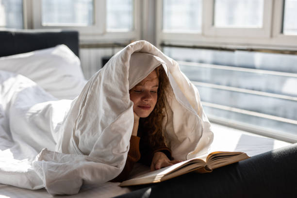 Charming young woman lying on bed and covering head with blanket