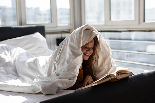 Charming young woman lying on bed and covering head with blanket