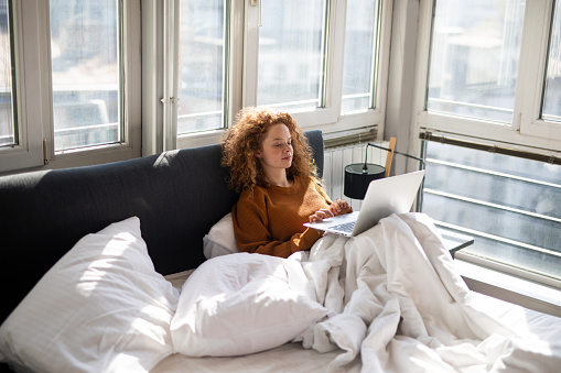 Young woman sitting on bed and using laptop