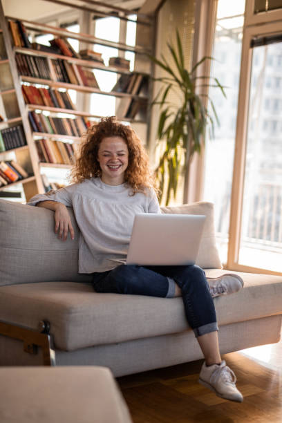 młoda kobieta siedząca na kanapie i korzystająca z laptopa - book sofa women bookshelf zdjęcia i obrazy z banku zdjęć