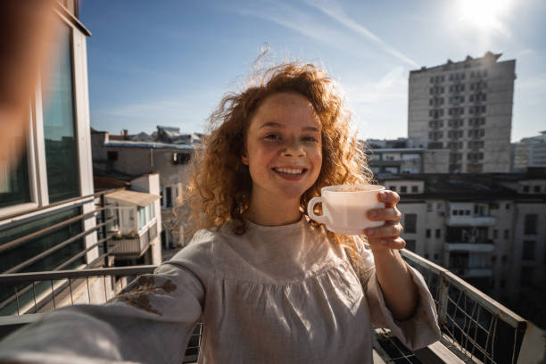 istock Happy young woman drinking coffee and taking selfie on balcony 1192478421