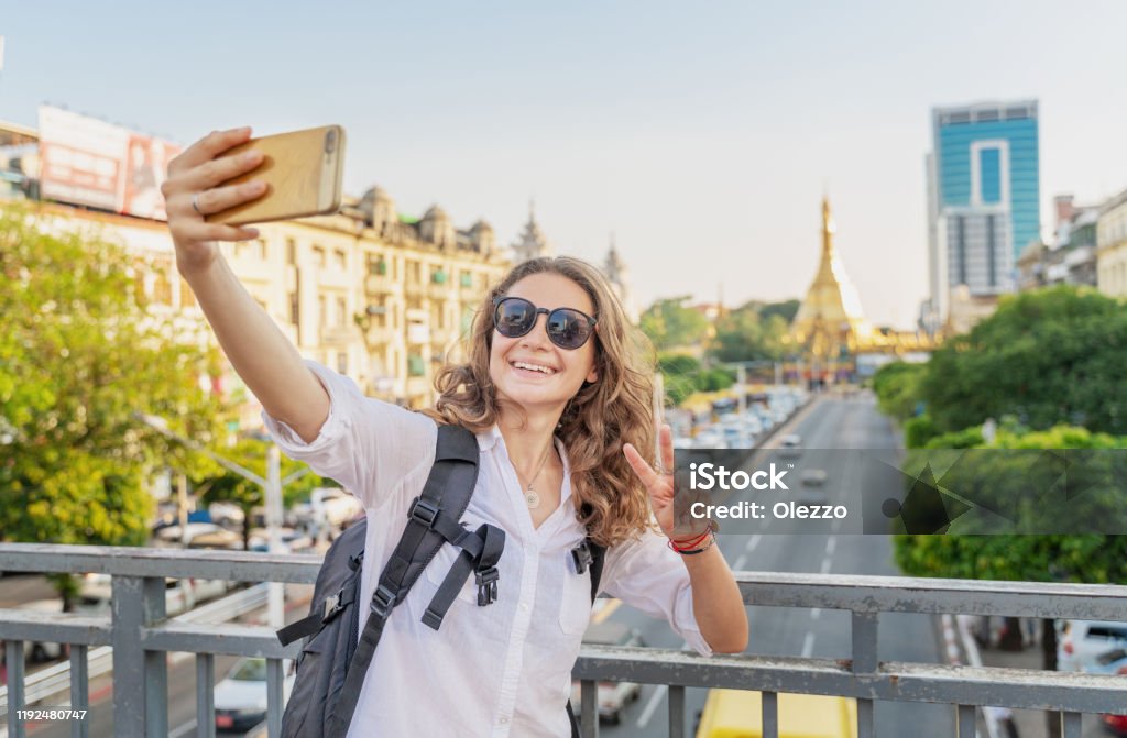 Young happy curly European girl traveler takes a selfie on the background of Yangon, the capital of Myanmar at Sule Pagoda Young happy curly European girl traveler takes a selfie on the background of Yangon, the capital of Myanmar at Sule Pagoda, Adult Stock Photo