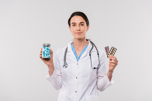 Pretty young female clinician in whitecoat holding jar with pills and blisters with painkillers in isolation
