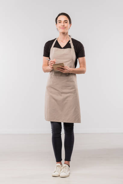 Pretty brunette waitress in workwear making notes in small notepad Pretty brunette waitress in workwear making notes in small notepad while standing in front of camera in isolation waitress stock pictures, royalty-free photos & images