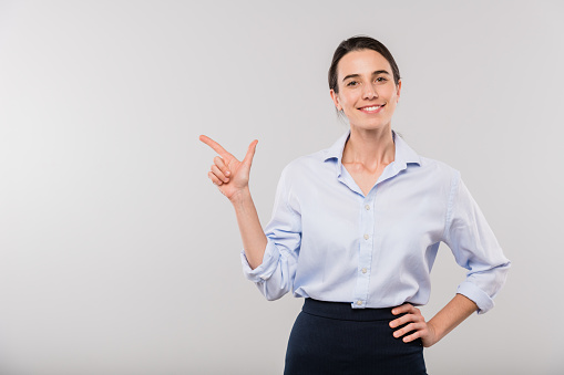 Happy young businesswoman in white shirt pointing aside while standing in front of camera in isolation