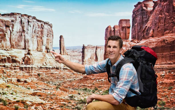 One man only / one person / waist up / side view / profile view of young men backpacker / tourist / tourist hiking / resting at the canyon / desert in front of landscape - scenery / nature / scenics - nature who by the sand who is outdoors wearing lumberjack shirt / plaid shirt / khaki pants / backpack who is smiling / happy / cheerful who is pointing and using hiking pole / sky / wild west / capitol reef national park / mountain / utah / wilderness area un hombre sólo / una persona / cintura para arriba / vista lateral / vista de perfil de los hombres jóvenes mochilero / turista / senderismo turístico / descansando en el cañón / desierto frente al paisaje - paisaje / naturaleza / escénicos - natural